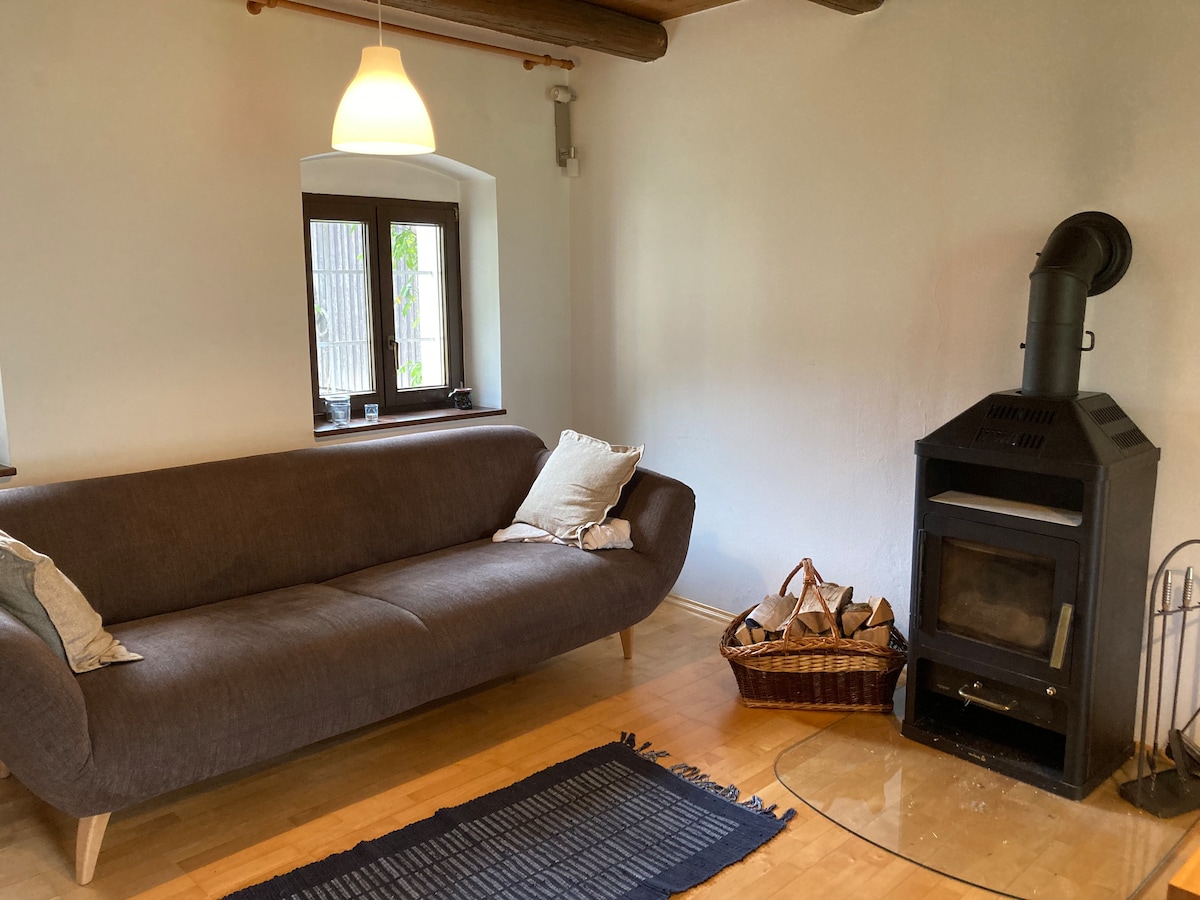 A cozy living area features a curved, dark upholstered sofa adorned with decorative cushions. A wood-burning stove stands adjacent, providing warmth. Natural light enters through two small windows. A woven basket beside the stove adds to the space's inviting atmosphere.