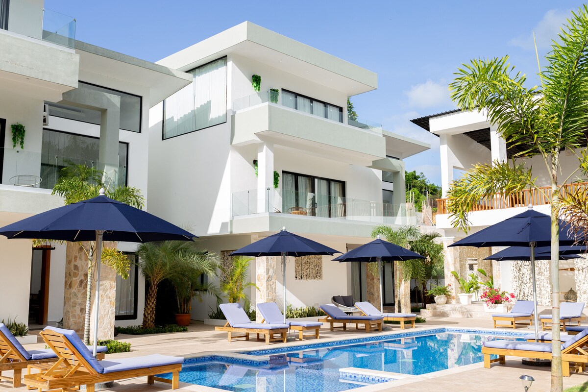 A modern villa is highlighted with a sparkling pool surrounded by sun loungers and large umbrellas. Lush tropical plants are positioned throughout the area, complementing the stylish architecture of the two-story building in the background.