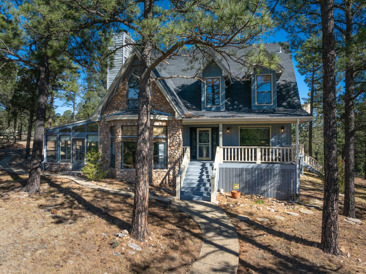 The exterior of a spacious home is surrounded by tall pine trees, featuring a charming stone facade and a sloped roof. A welcoming porch leads to the front door, while large windows offer glimpses into the interior. A pathway meanders through the natural landscape.