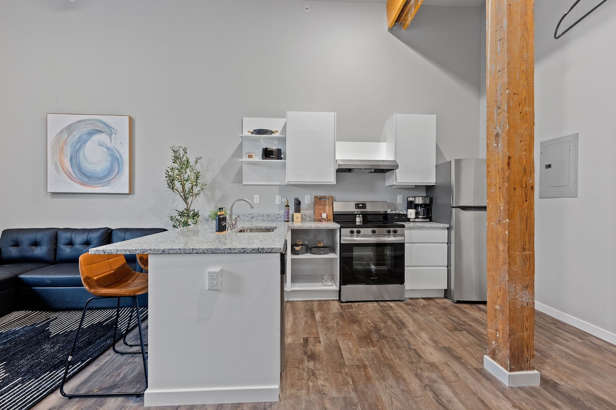 The modern kitchen showcases granite countertops, sleek cabinetry, and stainless steel appliances, including a stove and refrigerator. A spacious island with bar seating is positioned to one side, while a cozy seating area features a dark sofa, highlighting the open living concept.