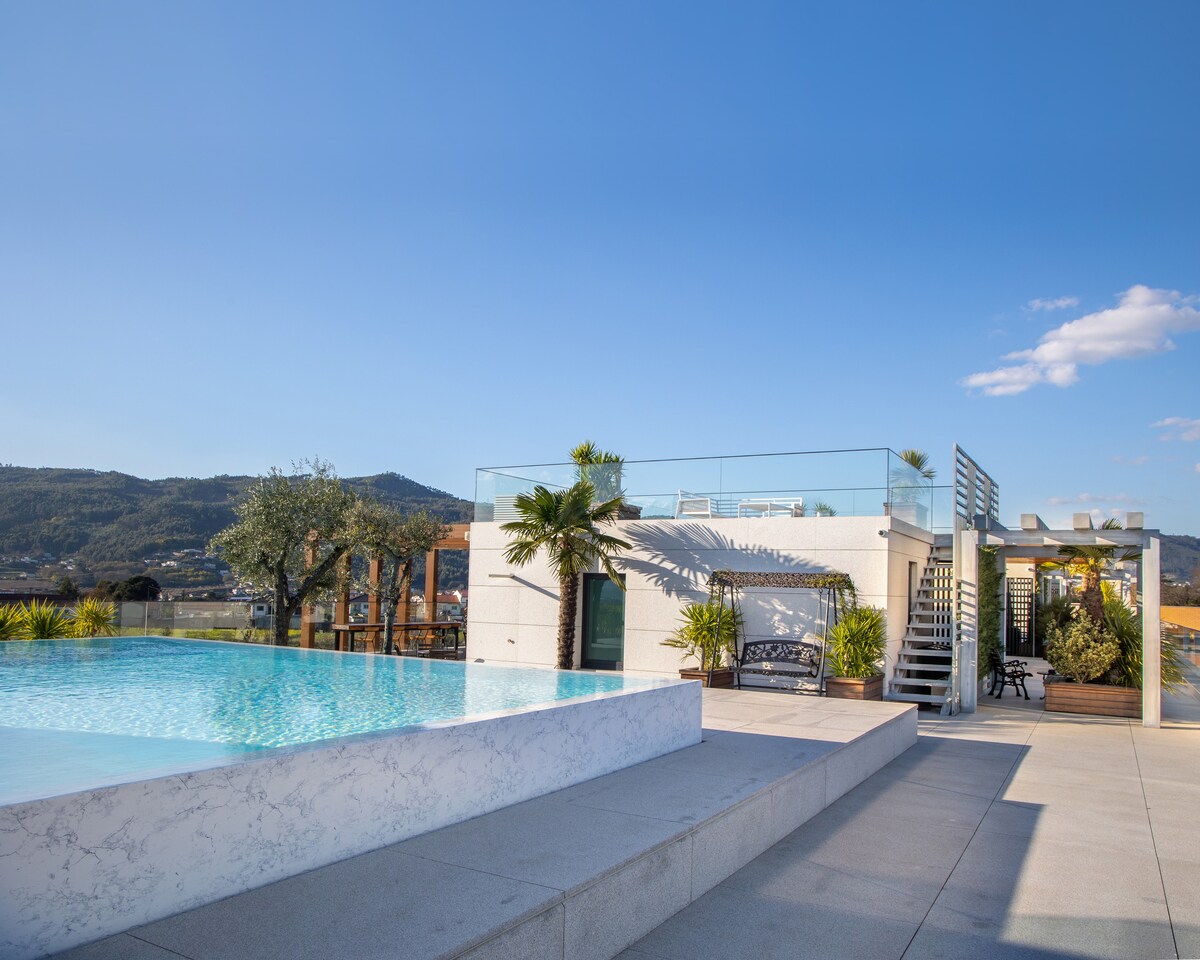 A modern outdoor space features a sleek infinity pool bordered by stone tiles. Lush greenery and palm trees are positioned around the pool area. A sun terrace with lounge seating is visible, along with a staircase leading to an upper deck with mountain views.
