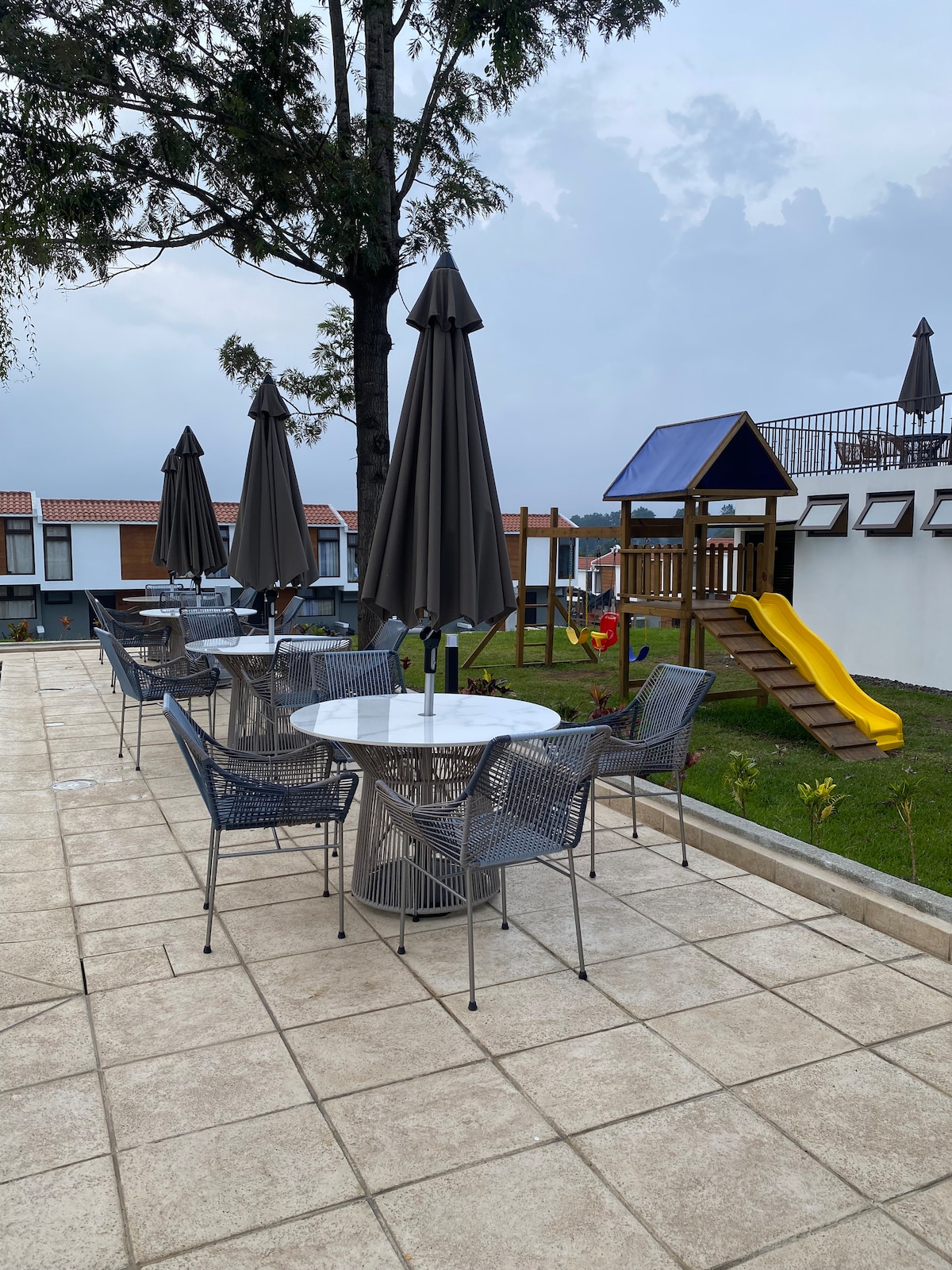 An outdoor area is showcased with several gray tables and chairs, accompanied by large umbrellas for shade. A playground is featured, complete with a slide and climbing structure, set against a backdrop of grass and a clear sky.