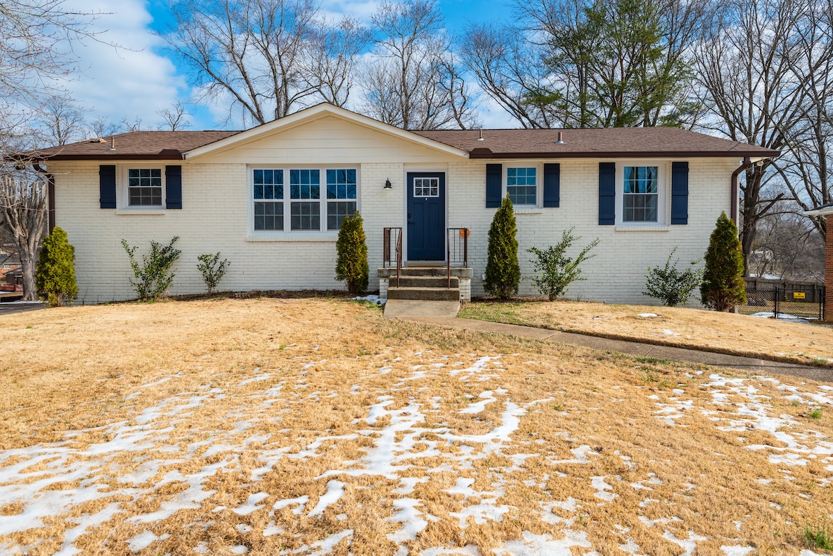 A single-story home with a white brick exterior is set against a clear blue sky. A neat lawn, partly covered with snow, frames the entrance. The home features two large front windows and a central door, flanked by shrubs and two small evergreen trees.