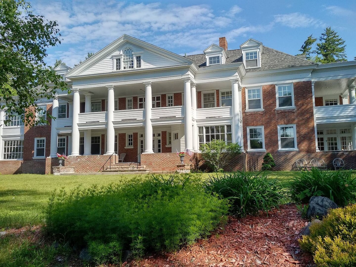 The grand facade of the Prescott House showcases classic architectural features, including large white columns and wide porches. Lush green lawns and well-maintained landscaping surround the brick structure, while a clear blue sky highlights the inviting atmosphere of this historic guesthouse.