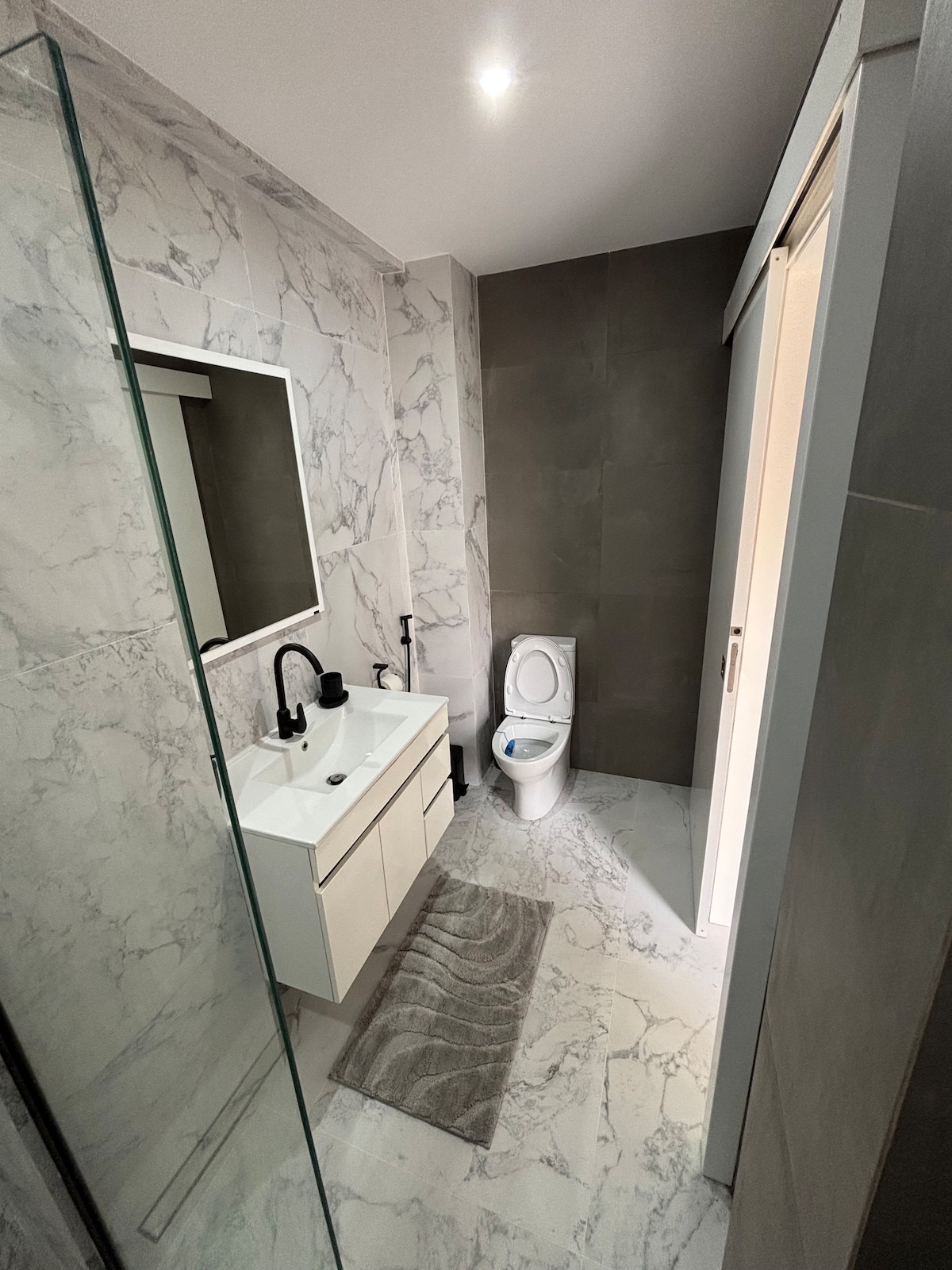 The modern bathroom features a walk-in shower with glass panels, a stylish sink with a mirror above, and a contemporary toilet. Gray and white marble tiles cover the walls and floor, providing a sleek appearance. A soft rug is positioned in front of the sink area.