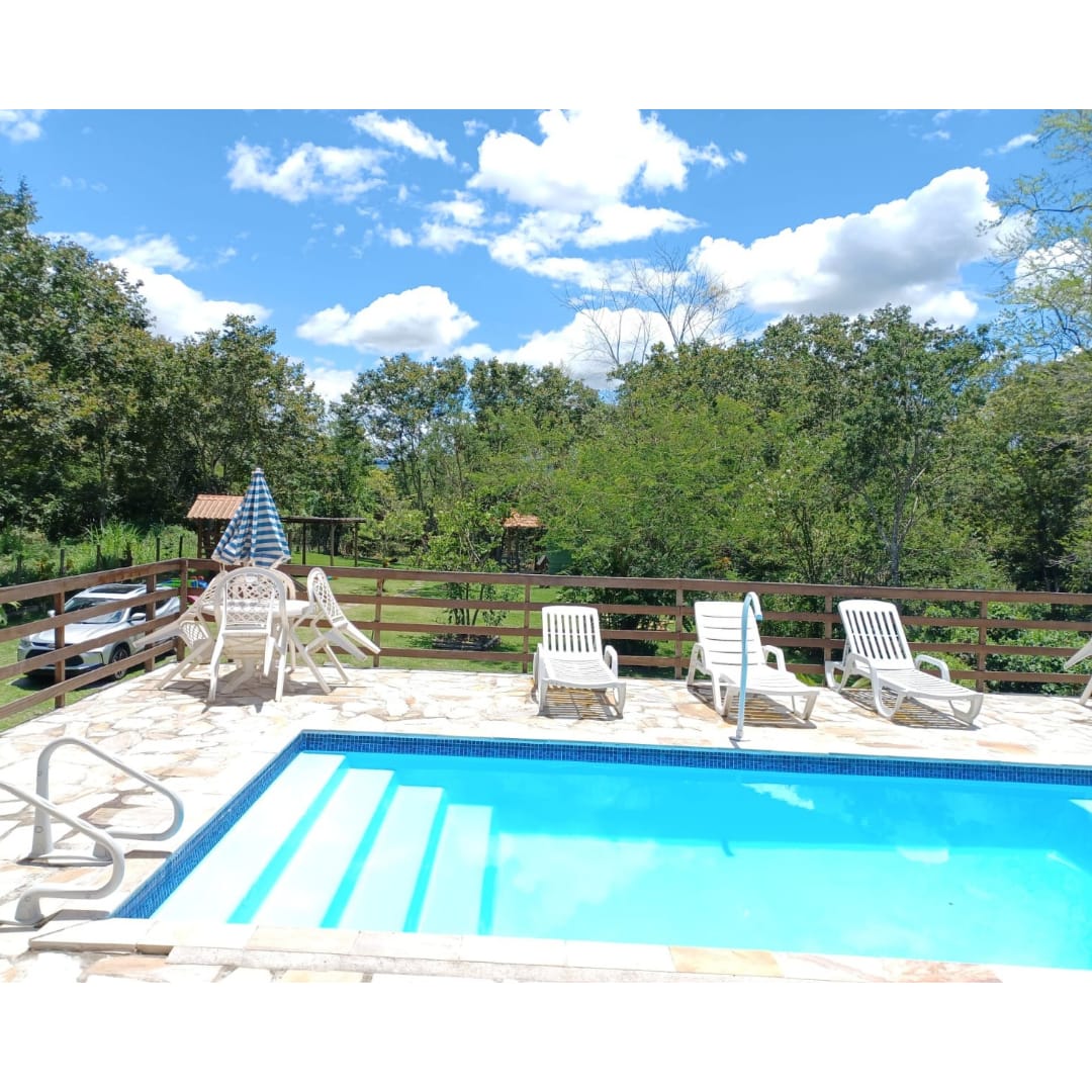 A serene pool area is framed by lush greenery and a wooden fence. Relaxing lounge chairs are positioned around the sparkling blue water. A shaded seating area is visible nearby, providing an inviting space for leisure under the sun.