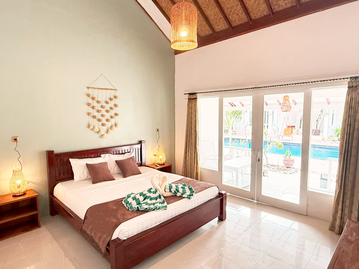 A spacious sleeping area features a wooden bed with neatly arranged linens and decorative towels. Soft lighting is provided by woven lamps on either side. Large glass doors open to a view of the pool area, allowing natural light to fill the room.