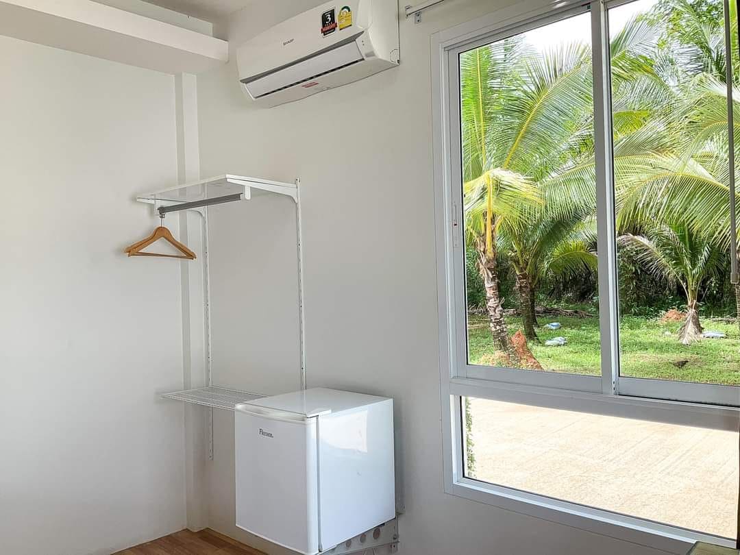 A bright room features a large window showcasing palm trees outside. An air conditioning unit is installed above a compact refrigerator. A hanging rack with a single wooden hanger is mounted on the wall, providing additional storage options.