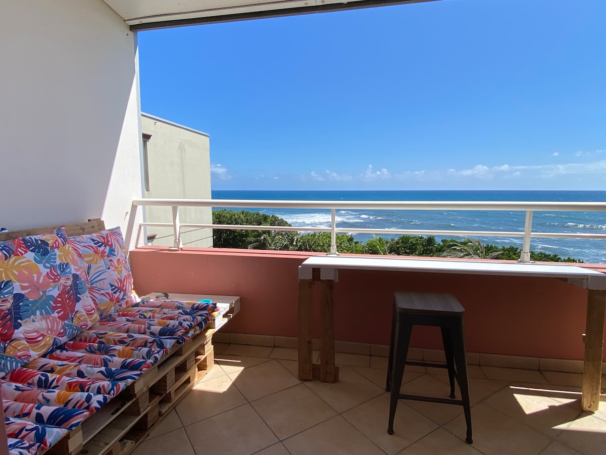 A spacious outdoor terrace is equipped with a colorful cushioned seating area made from wooden pallets. A bar-style table is positioned next to the seating, offering a clear view of the coastline and blue ocean under a bright sky.