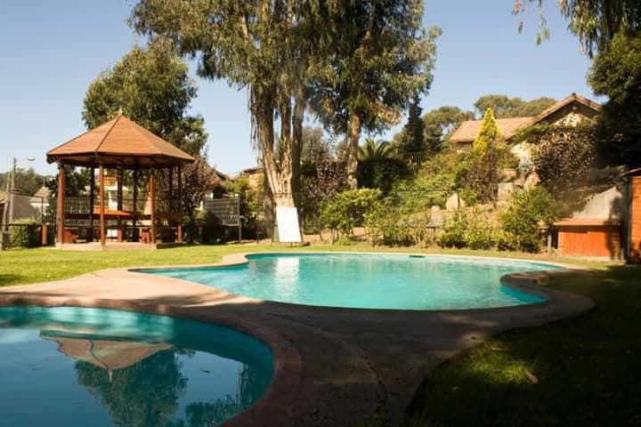 An outdoor pool is framed by lush green grass and surrounded by trees. A gazebo with a wooden table and benches sits nearby, providing a shaded area for relaxation. The landscape includes well-maintained gardens, enhancing the tranquil setting.
