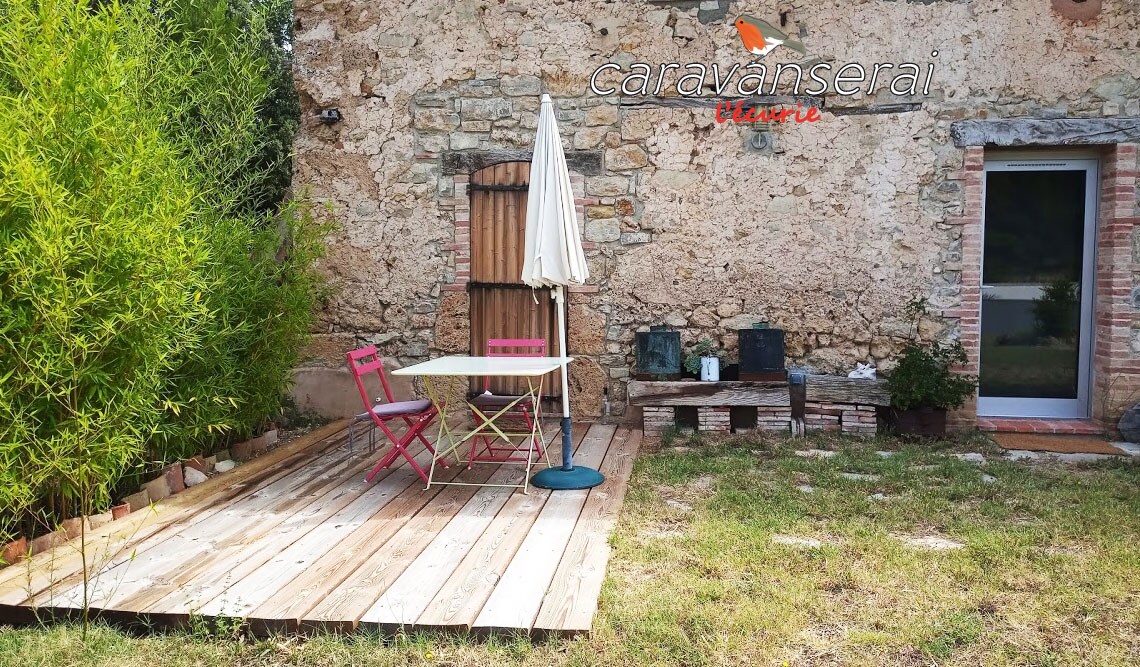 A wooden terrace is presented, featuring a round table with two colorful chairs and an umbrella. The setting is surrounded by greenery and the rustic stone wall of the accommodation can be seen in the backdrop.
