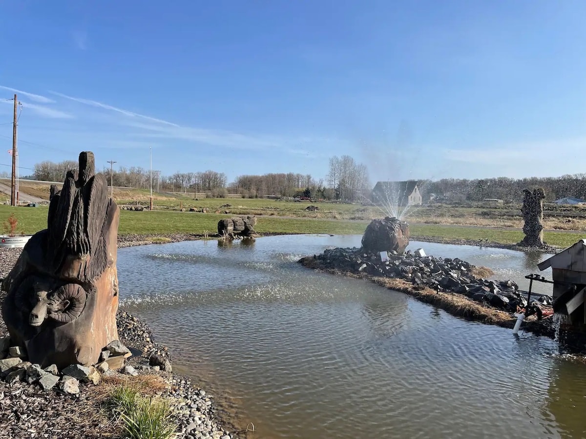 A serene pond is framed by natural landscaping, featuring two stone sculptures and a gentle fountain. Water bubbles up from the fountain, creating a peaceful ambiance. The surrounding fields and trees can be seen in the distance, adding to the tranquil setting.