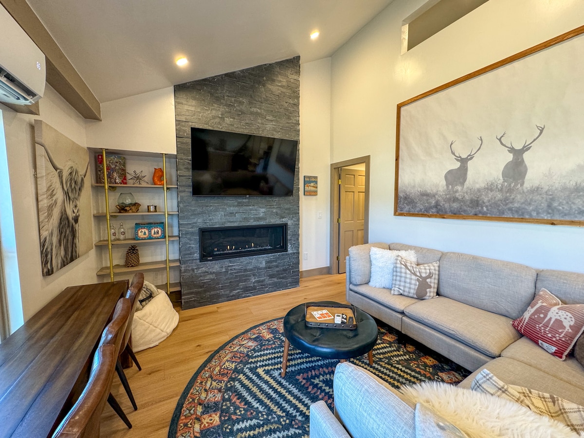 A welcoming living area features a modern gas fireplace set against a stone wall, with a large flat-screen TV mounted above. A sectional sofa is arranged around a circular coffee table on a patterned rug. A dining table is visible, accompanied by decorative shelves.