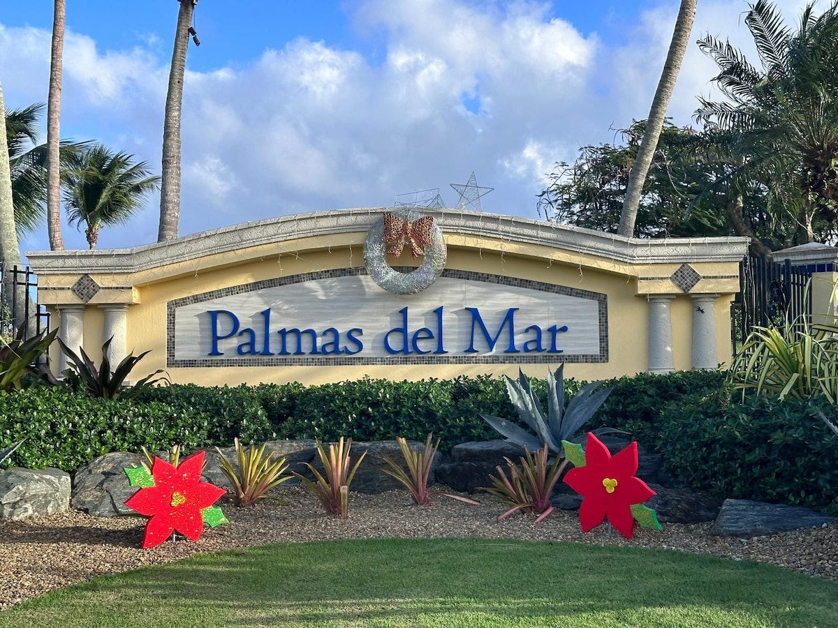 The entrance sign of Palmas del Mar is prominently displayed, featuring elegant lettering against a cream-colored backdrop. Festive holiday decorations, including wreaths and floral arrangements, are positioned in the foreground, surrounded by lush greenery and palm trees under a partly cloudy sky.