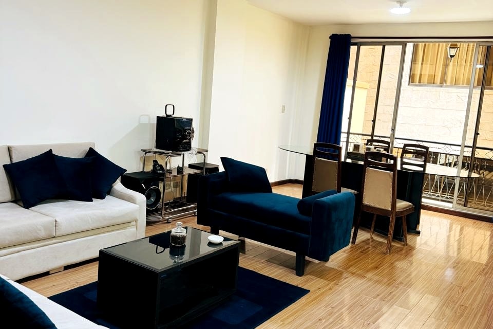 A spacious living area features a light-colored sofa and a dark blue armchair, creating a balanced decor. A black coffee table rests on a light-colored rug, and a glass dining table with chairs is visible in the background, accompanied by large windows presenting views of the outdoor space.