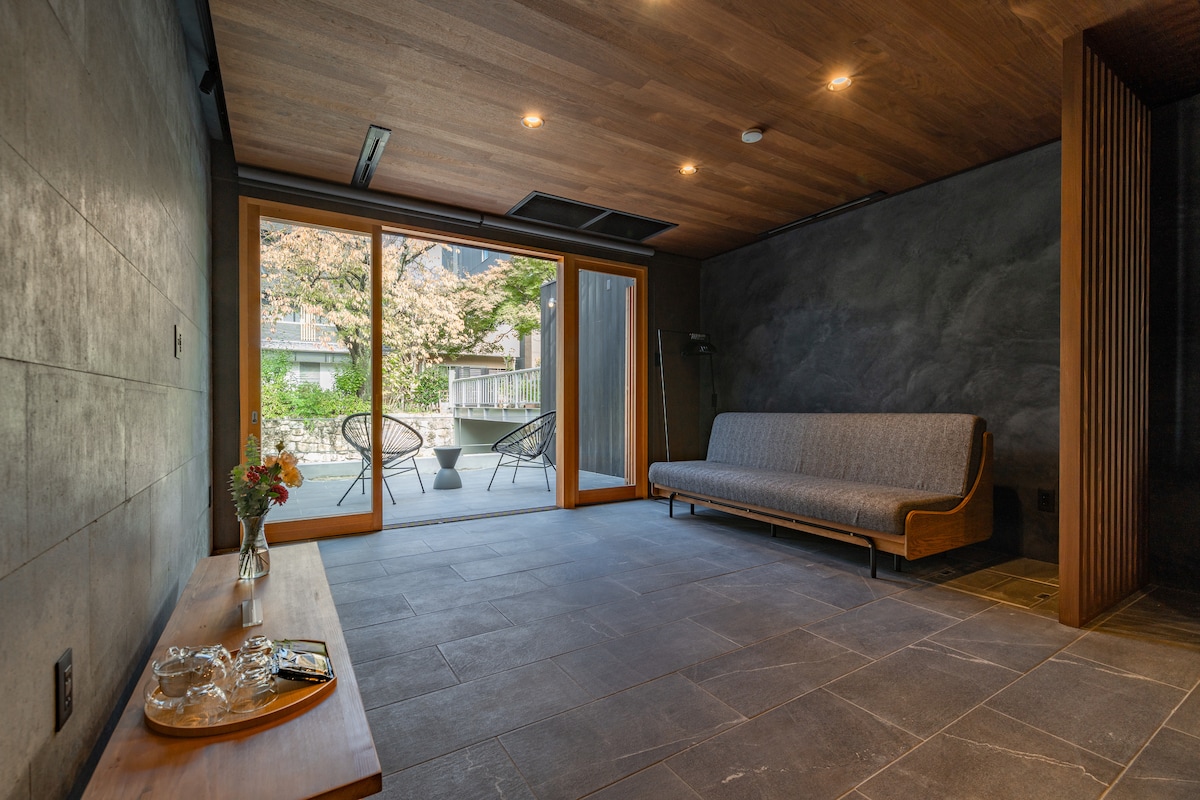 A serene living area is highlighted by a smooth stone floor and wooden ceiling. Natural light enters through large glass doors, which open to an outdoor view. A simple wooden table displays a vase of flowers, while a gray sofa offers comfortable seating.