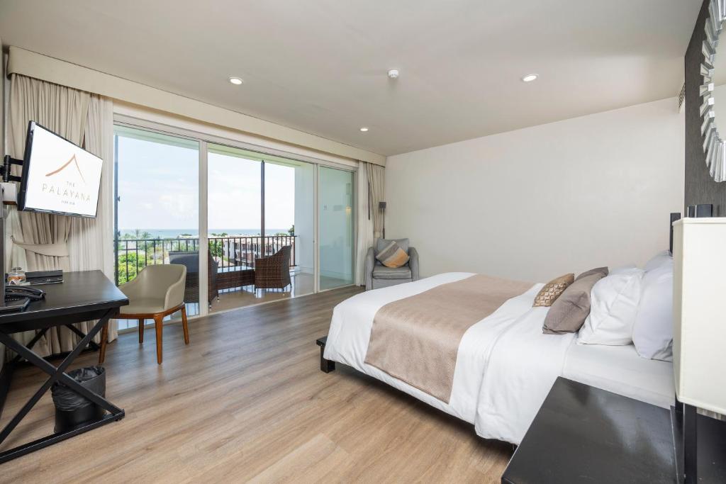 A spacious room is presented with a large double bed and a modern desk. Natural light filters through large sliding glass doors that lead to a private balcony, which includes a seating area. Soft curtains complement the neutral-toned decor and wooden flooring.