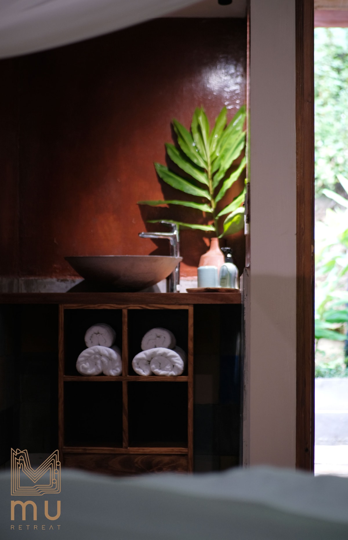 A sleek wooden vanity showcases a round vessel sink, accompanied by a small plant and decorative items. Neatly stacked towels are stored within an open shelf, enhancing the organized and serene environment. Natural light filters through a doorway, illuminating the space.