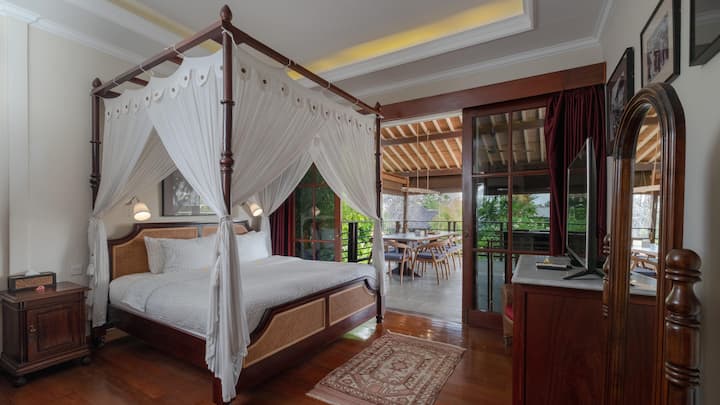 A charming colonial-style room with a majestic four-poster bed, classic wooden furnishings, and an airy balcony that opens to lush greenery, offering a tranquil and timeless retreat.