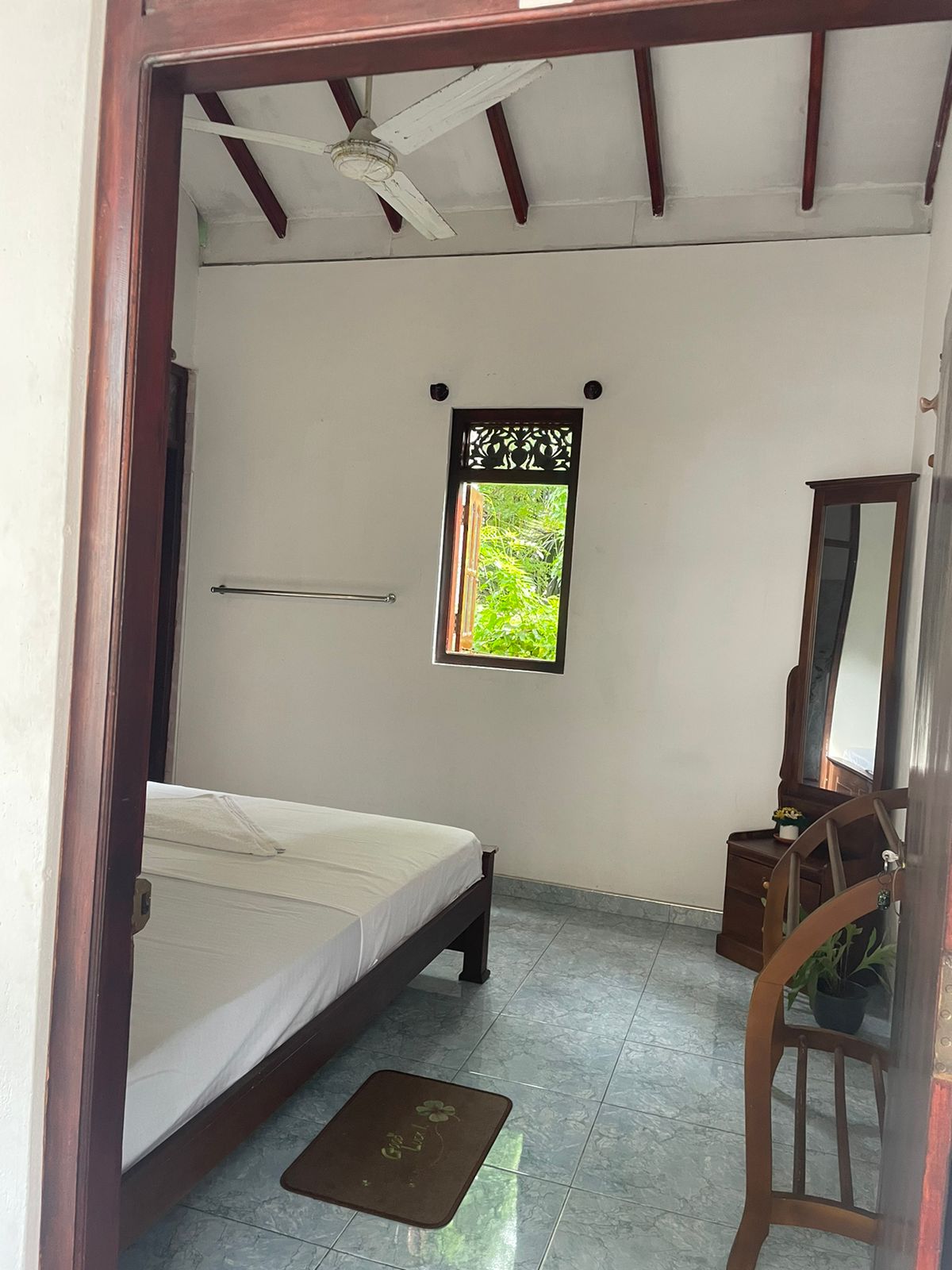 A simple sleeping area is seen, featuring a spacious bed with white linens. A wooden mirror and chair are positioned against one wall. A small window allows natural light to enter, framed by intricate woodwork, while a light-colored rug lies on the tiled floor.