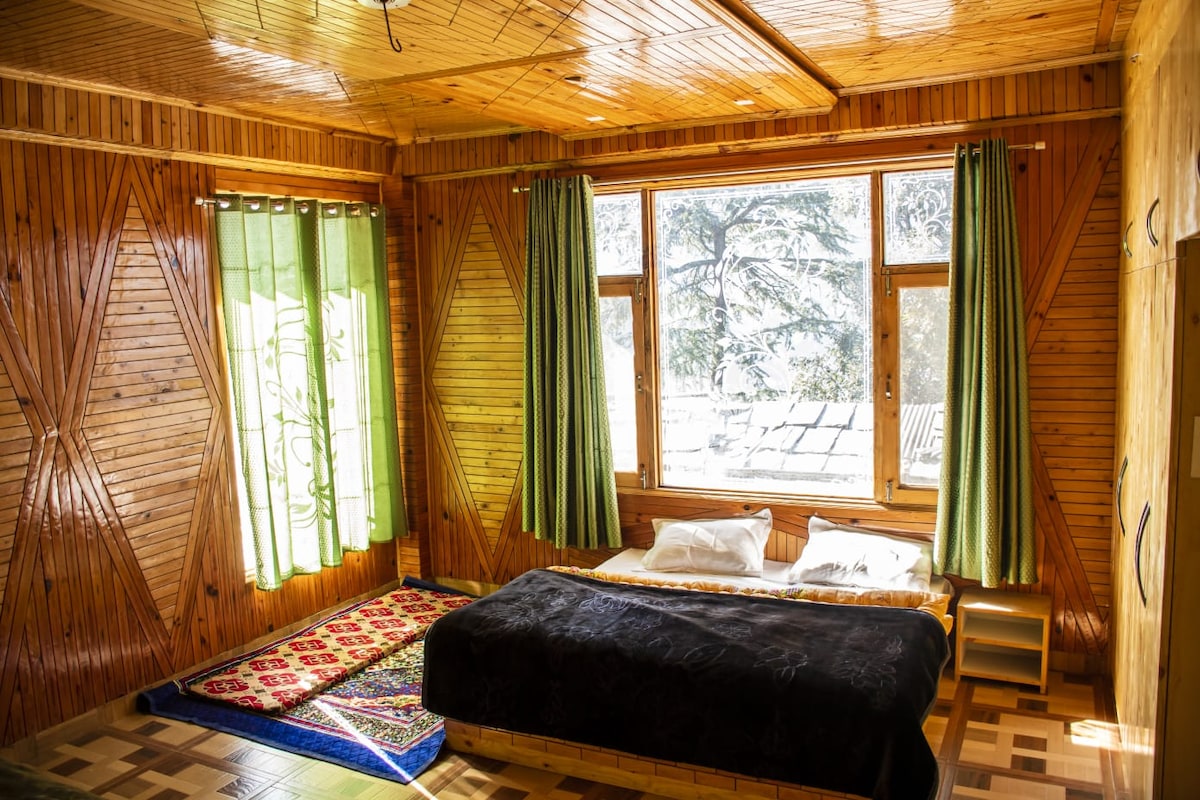 A spacious room features wooden paneling and large windows dressed with green curtains, allowing natural light to fill the space. A black blanket is laid over the bed, complemented by white pillows. A decorative rug adds color to the floor beside the bed.