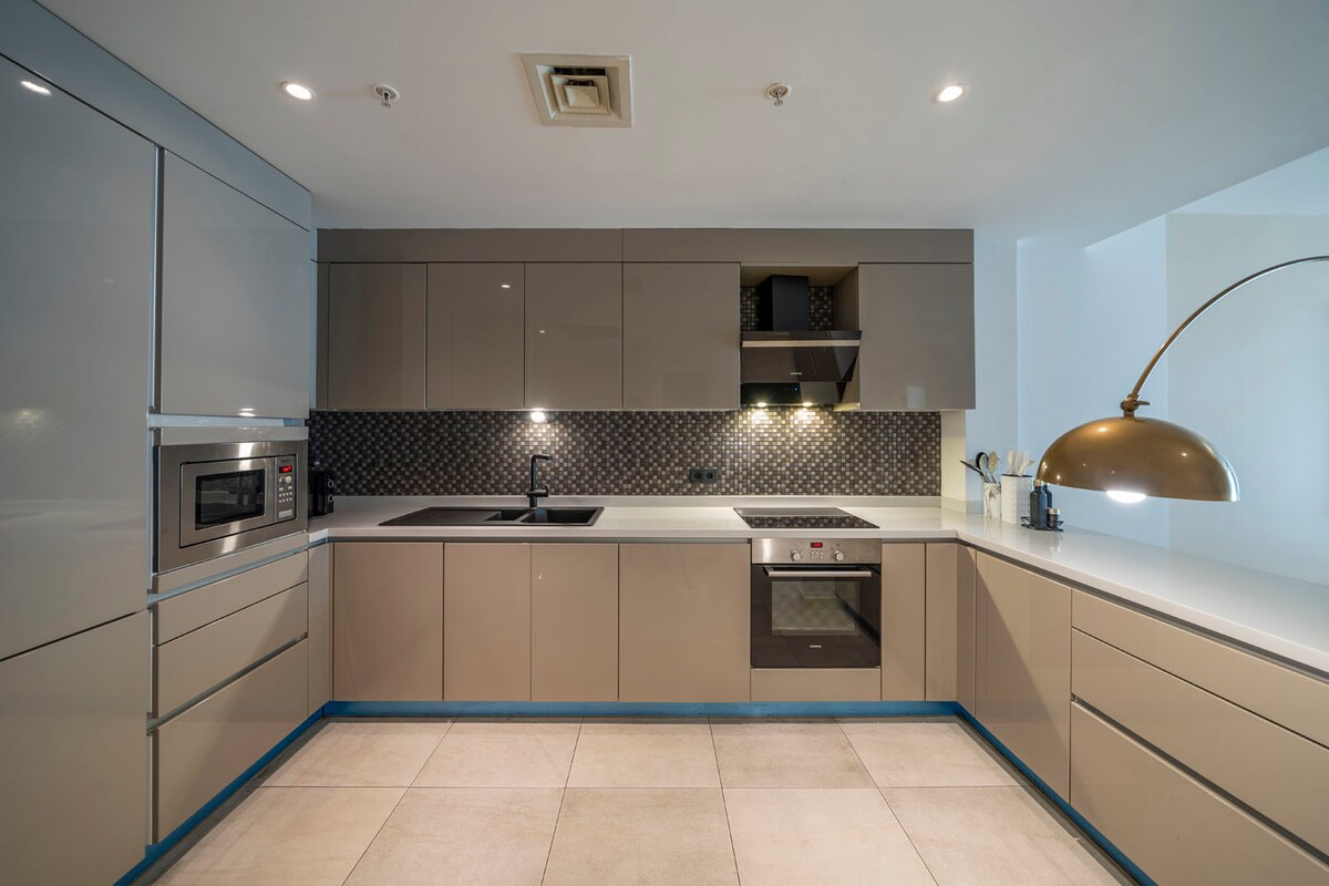 A modern kitchen is equipped with sleek cabinetry in neutral tones. A built-in microwave and oven, along with a stovetop, are visible. A large gold pendant lamp hangs above the counter, which features a subtle textured backsplash. Light-colored tiles cover the floor.