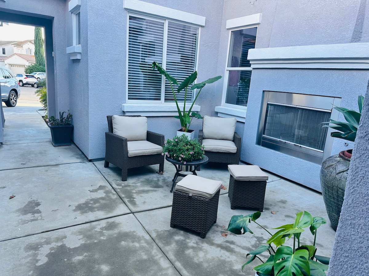 An outdoor seating area is set on a concrete pathway, featuring a round table surrounded by four wicker chairs with light cushions. A potted plant with large green leaves is present, and a wall-mounted fireplace adds a modern touch. Natural light filters through the nearby windows.
