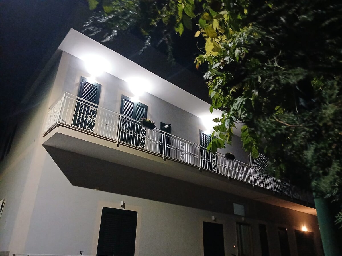 The structure is illuminated under a night sky, showcasing a modern two-story façade with a balcony adorned with potted plants. The exterior features contrasting light and shadow, highlighting its fresh architectural lines.