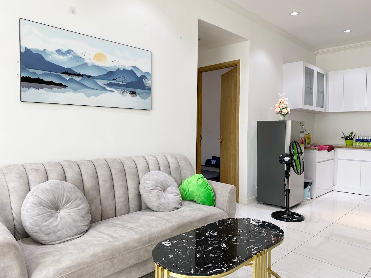 A light-filled living area features a plush gray sofa adorned with decorative pillows in various shades. A marble-top coffee table is positioned in front of the sofa. The background reveals an open kitchen with modern cabinetry and a small dining area.
