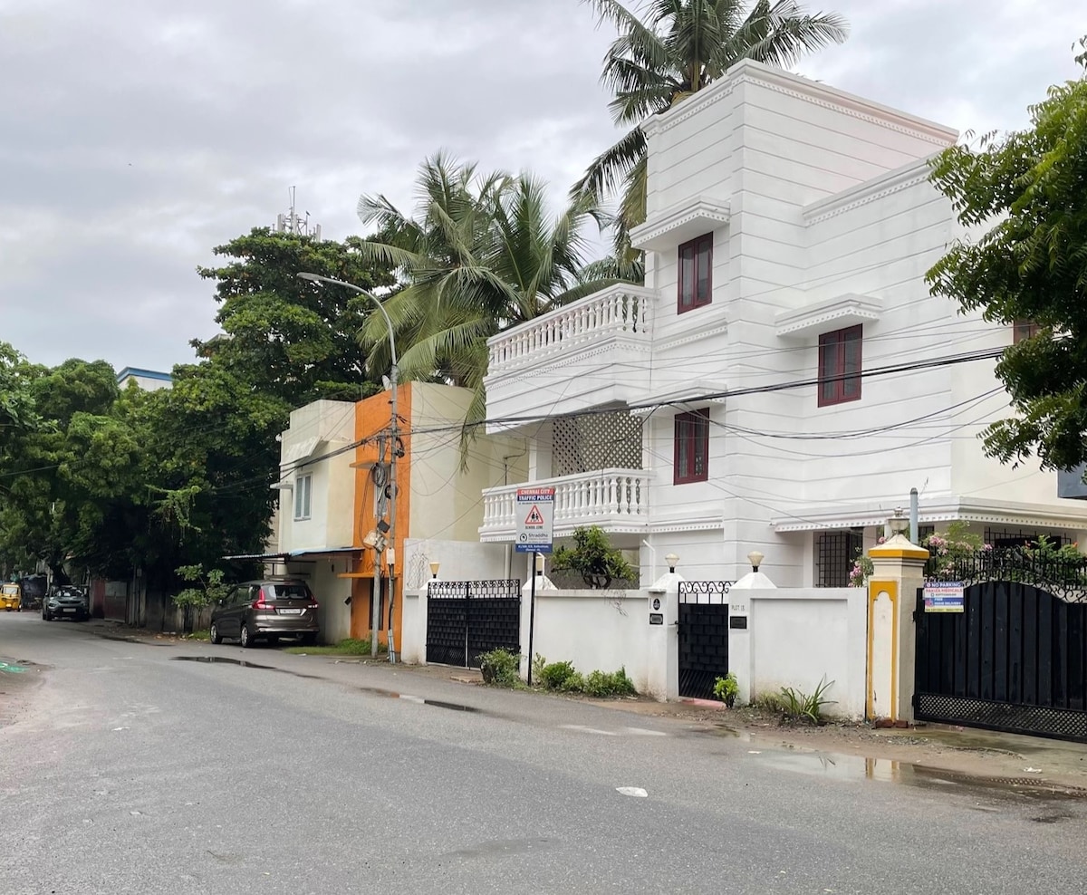 The exterior of a two-story white building is displayed, featuring a balcony and intricate railings. Surrounding greenery includes palm trees, while the street is lined with parked vehicles and a neighboring orange structure. A clear sky indicates overcast weather.