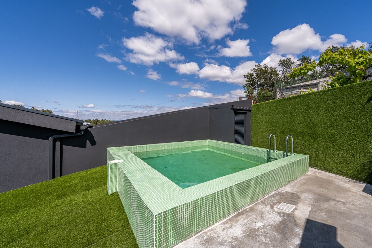 A rectangular pool with a green-tinted water surface is surrounded by a textured, grassy area. The pool is framed by a low, transparent wall. The backdrop features a clear blue sky and scattered clouds.