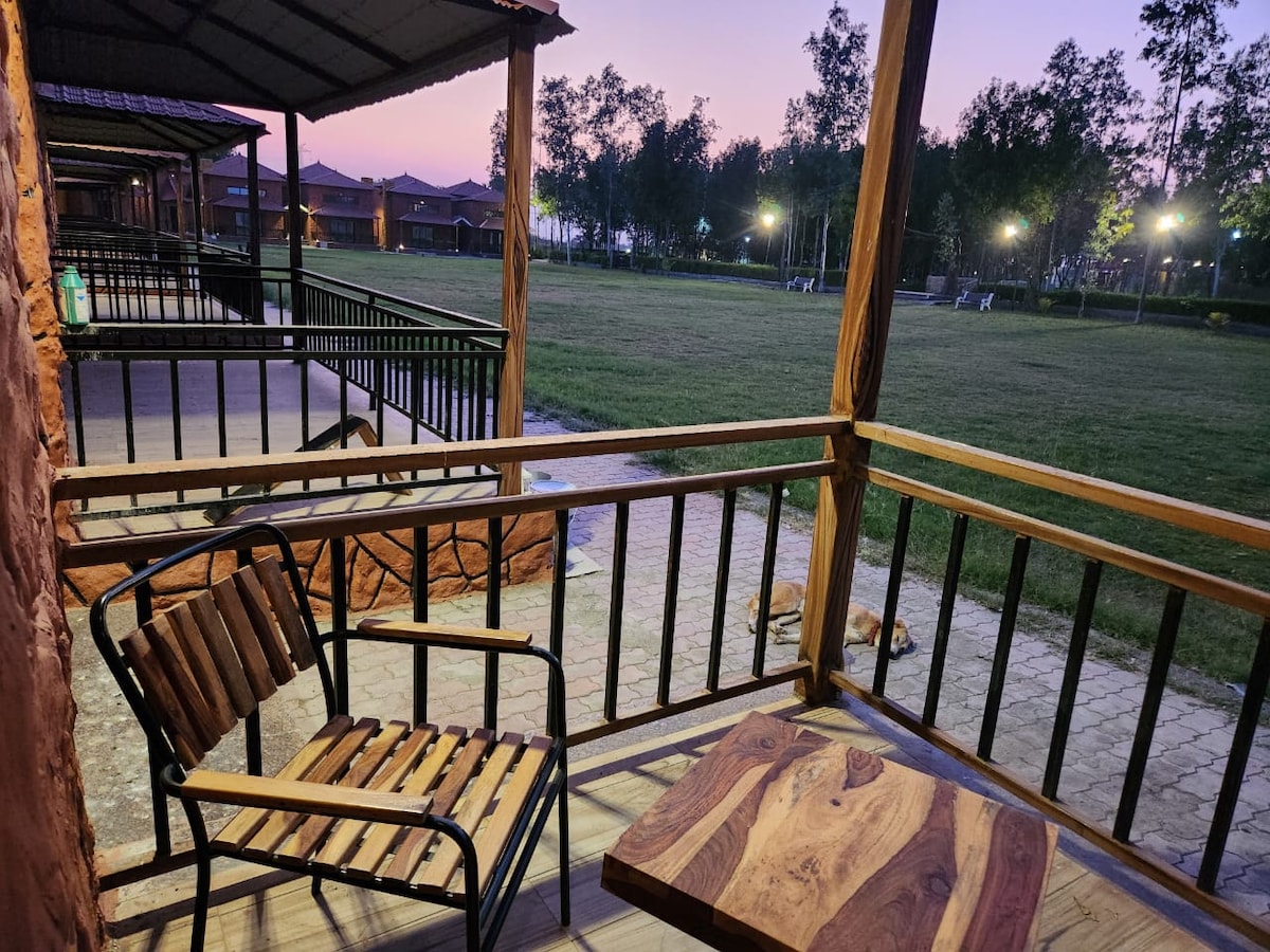A covered porch is visible, featuring a wooden table and two chairs. The surrounding area includes a grassy field and trees, with pathway lights gently illuminating the scene as twilight settles in.