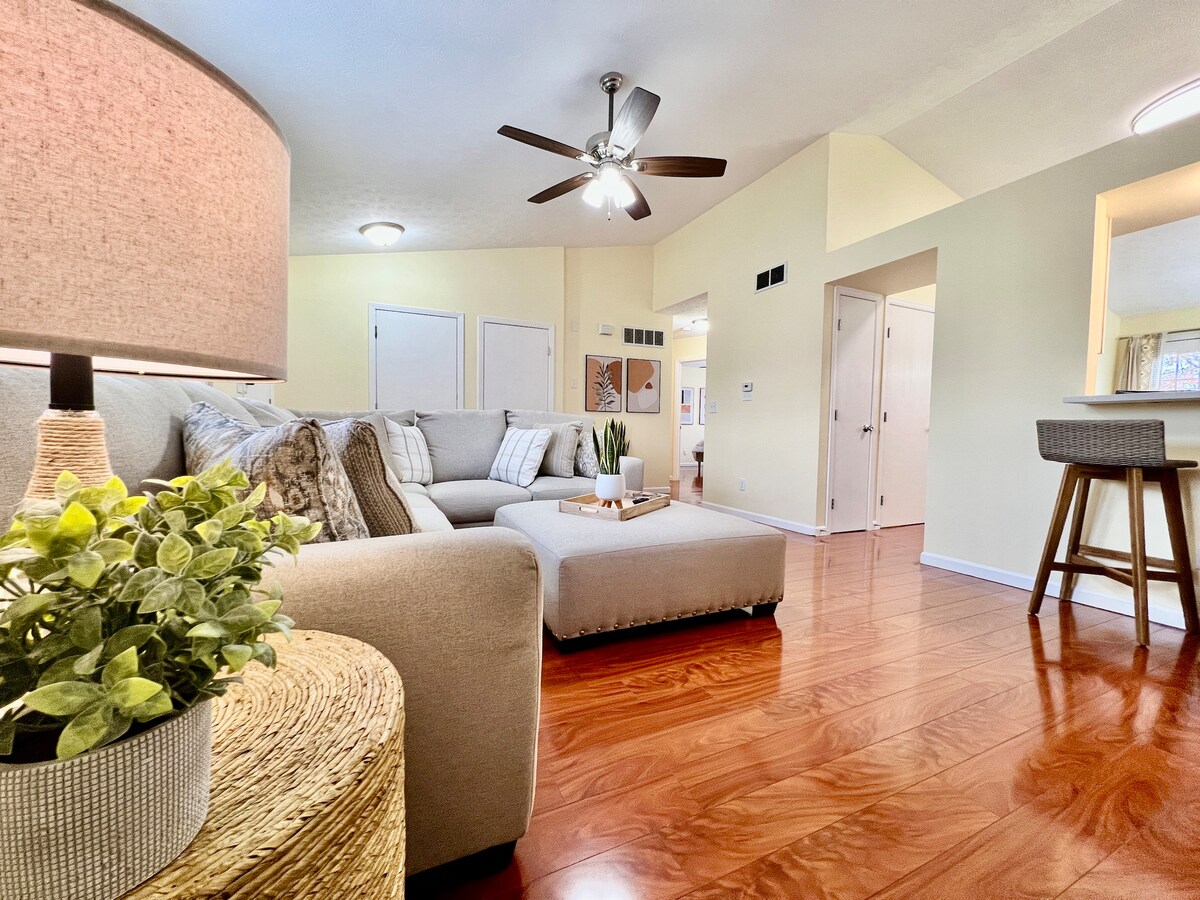 A cozy living area is presented with a sectional sofa, a stylish lamp, and a decorative plant. Light hardwood flooring complements the neutral walls and ceiling fan, creating an inviting space that flows into the adjacent dining area and kitchen.