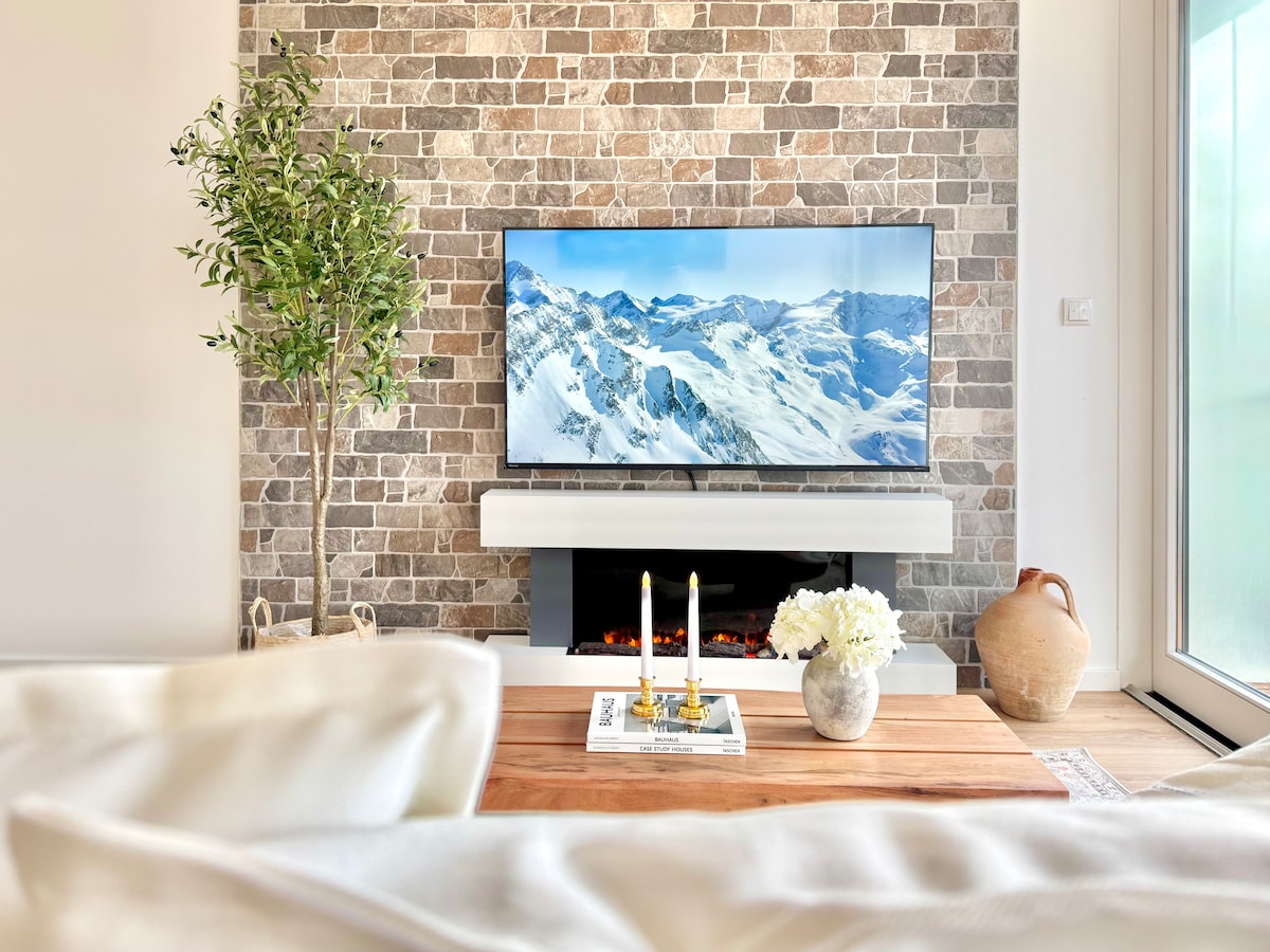 A spacious living area is showcased, featuring a modern stone accent wall and a flat-screen television displaying snowy mountains. A coffee table with candles and a decorative vase enhances the space, complemented by a small indoor plant. Light streams in from a large window.