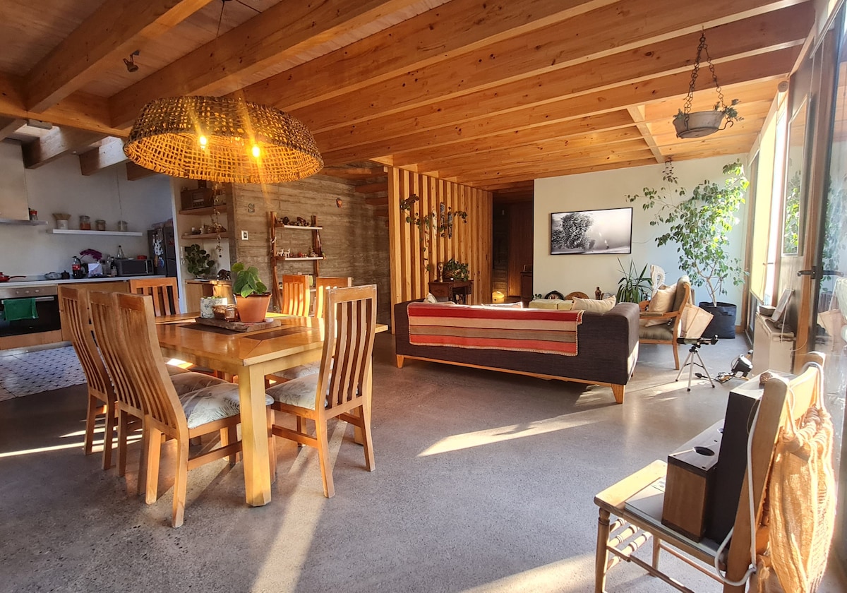 A spacious living and dining area features a wooden dining table surrounded by chairs. Natural light streams through large windows, illuminating the wooden ceiling beams and various plants. A cozy seating area includes a sofa and accent chairs, creating an inviting space for relaxation.