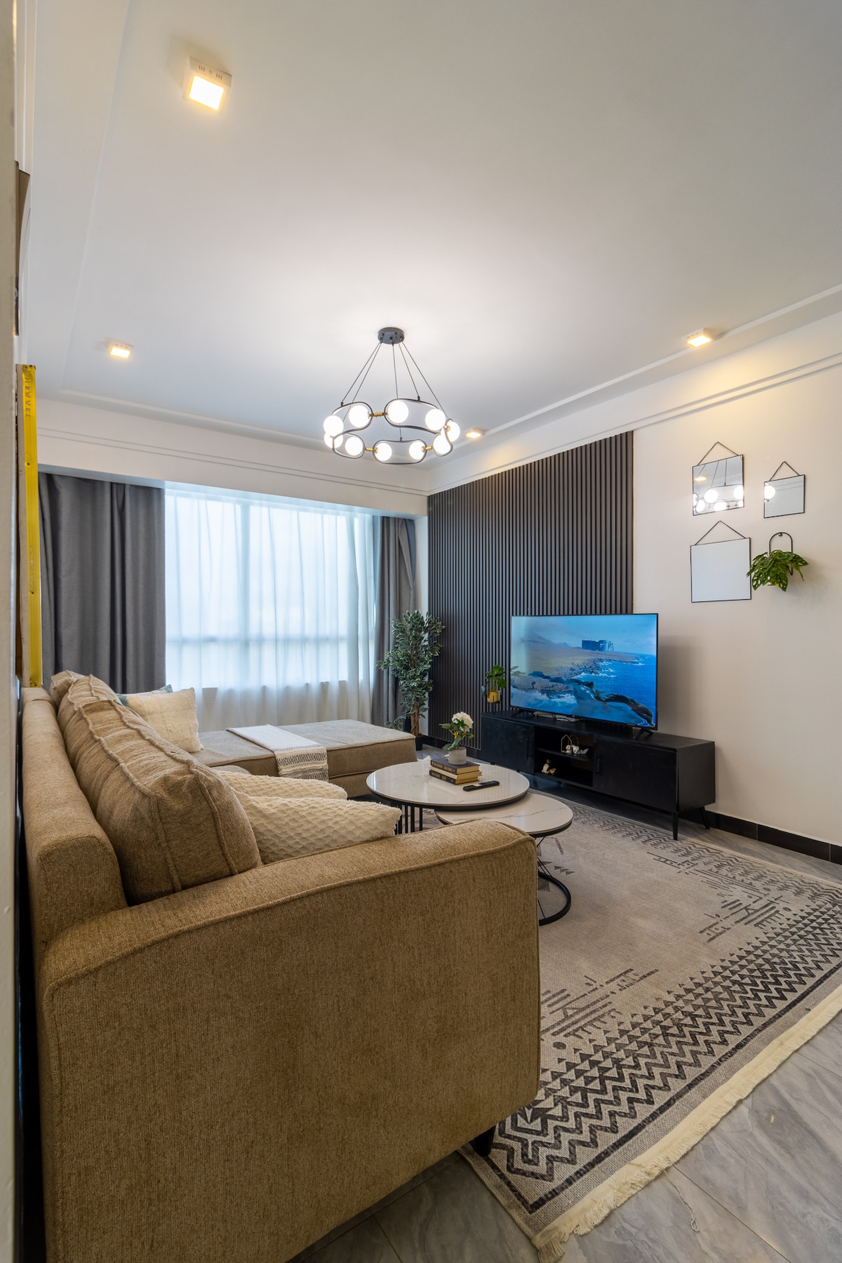 A cozy living area features a plush sectional sofa arranged around a circular coffee table. A television is mounted on the wall, while light filters through sheer curtains. Decorative plants and geometric wall art add a touch of modern elegance to the space.