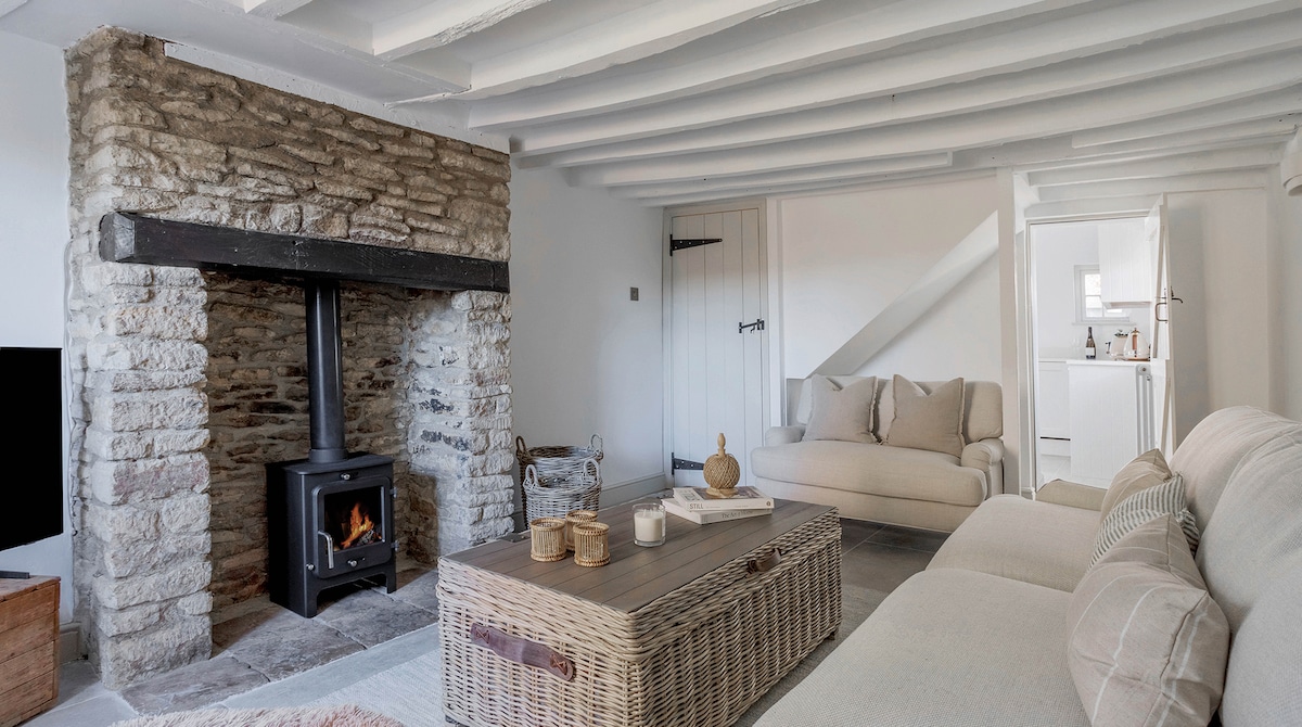 A spacious living area is highlighted by a rustic stone fireplace, featuring a wood-burning stove. A comfortable sofa and a stylish coffee table made of natural materials are arranged to create an inviting space. Light filters through the room, enhancing the overall ambiance.