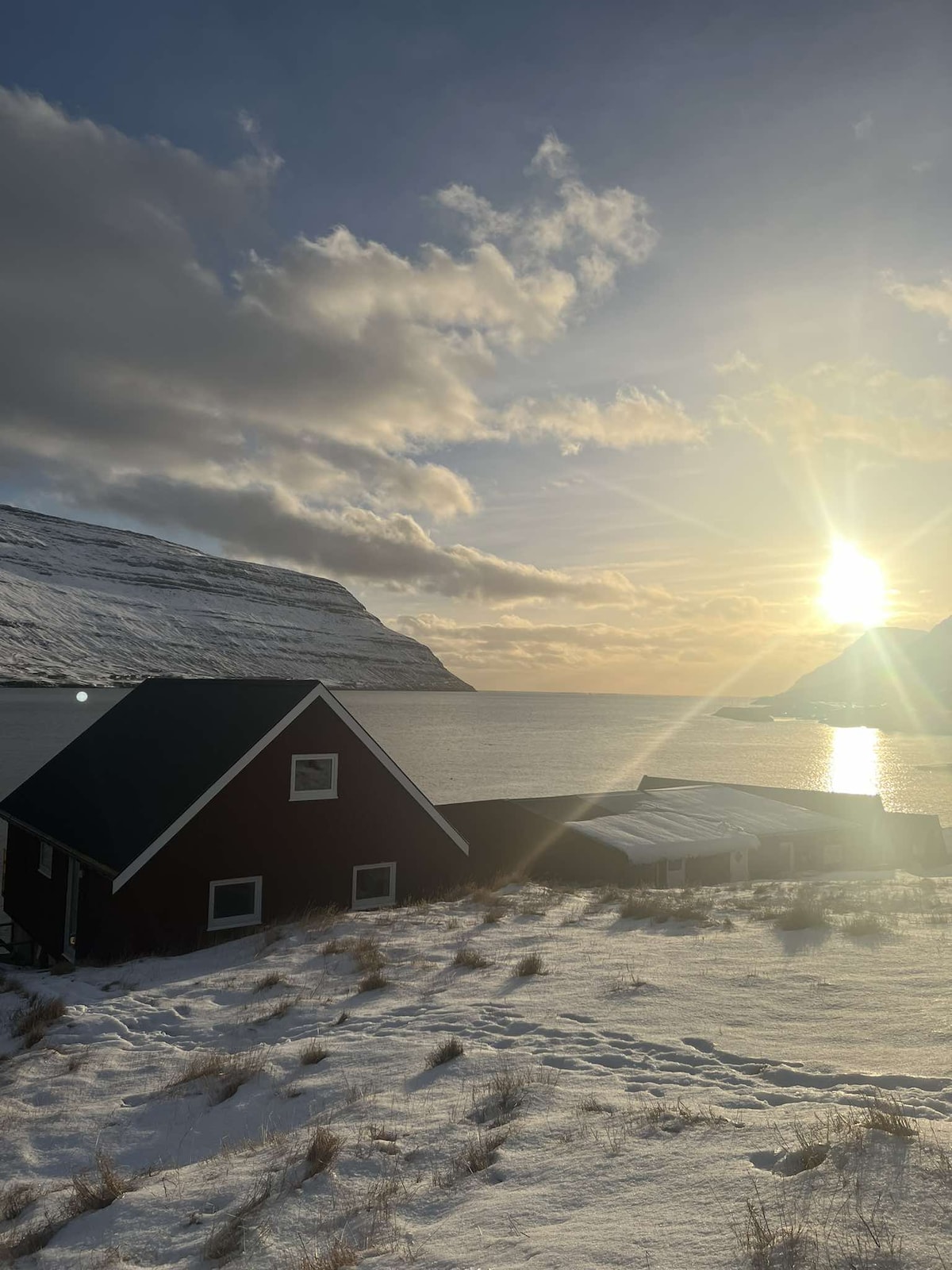 A red cabin is positioned near a tranquil body of water, with snow-covered ground surrounding it. The sun rises above the mountains in the background, casting a gentle glow on the landscape, while clouds drift across a brightening sky.