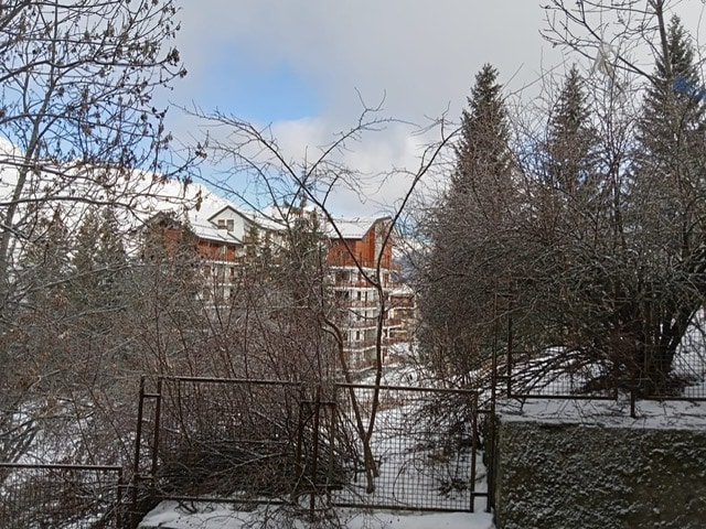 A winter landscape is presented, featuring a view of a multi-story building surrounded by snow-covered trees and shrubs. The scene is complemented by a partly cloudy sky, enhancing the tranquil ambiance of the setting.
