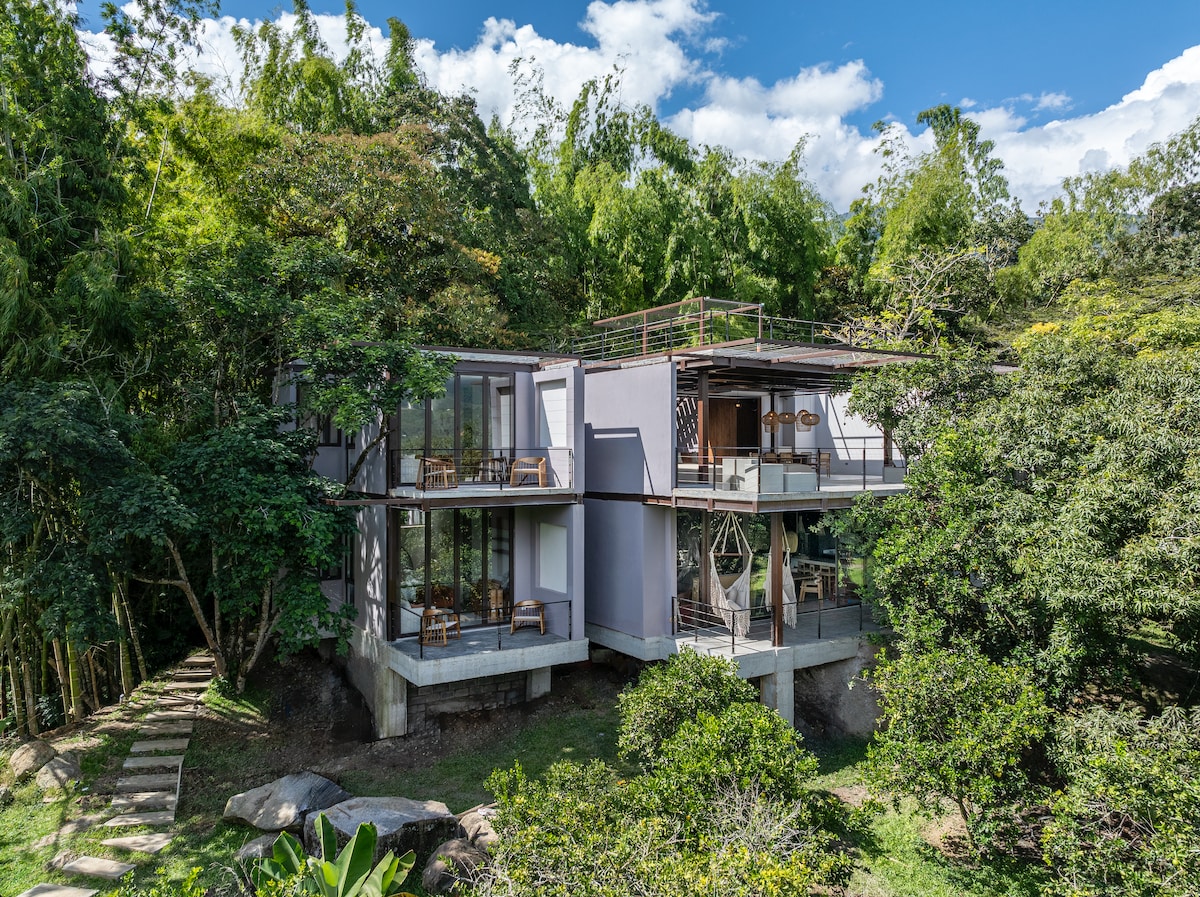 The modern house sits amidst lush greenery, showcasing a multi-level structure with expansive glass windows. The outdoor terrace features seating areas and is surrounded by trees, enhancing the connection with nature. Natural light fills the open spaces, providing an inviting atmosphere.
