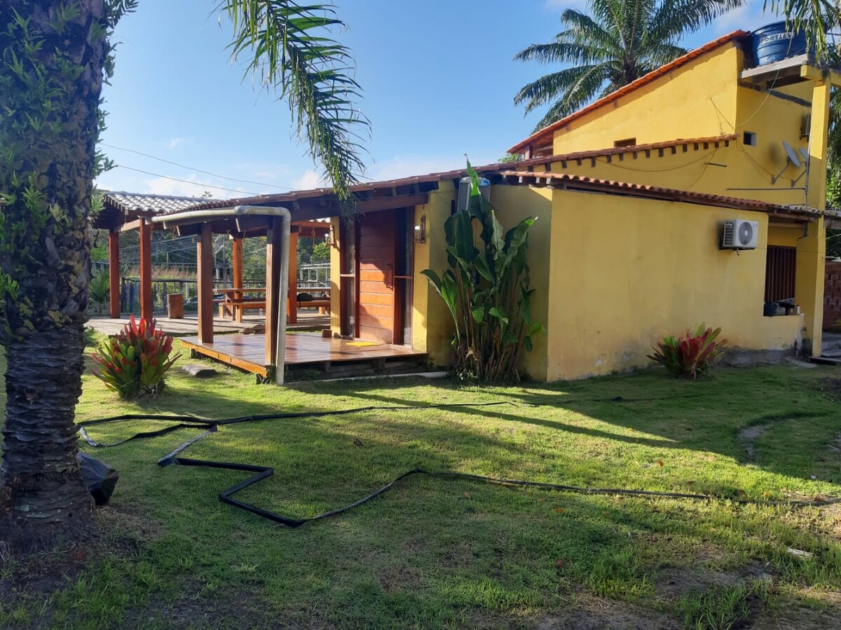 A charming exterior features a vibrant yellow house surrounded by lush greenery and palm trees. A wooden patio is visible, enhancing the outdoor space, while the air-conditioning unit is mounted beside a wall. Sunlight casts gentle shadows on the well-maintained grass.
