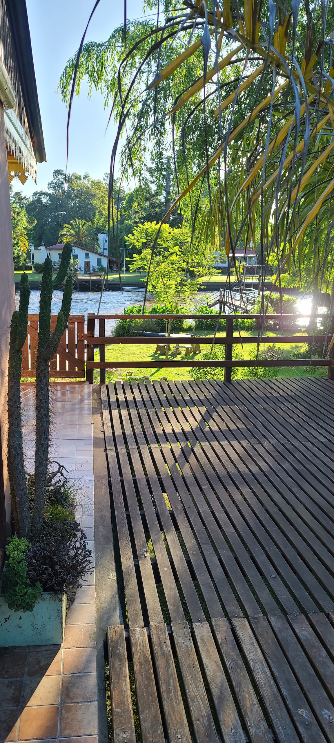 A sunlit outdoor deck features wooden planks, providing a spacious area for relaxation. Lush green plants and a cactus are visible, enhancing the natural scenery. The tranquil view of a waterway and surrounding greenery is framed by fencing, inviting peaceful moments in nature.