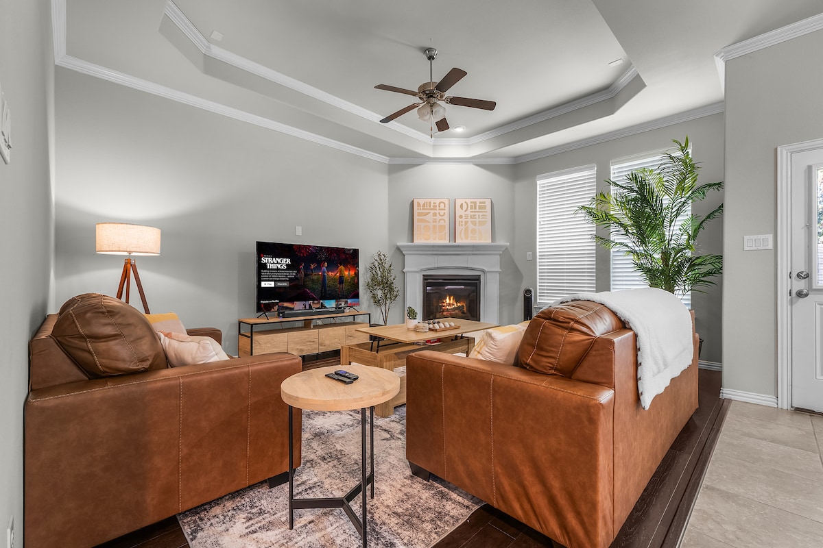 A spacious living area is highlighted by tan leather sofas positioned around a wooden coffee table. A large television is mounted on the wall opposite a cozy fireplace, with soft light filtering through sheer window coverings. Decorative plants add a touch of greenery.