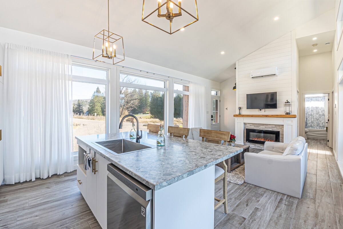 An open-concept living space is shown, featuring a modern kitchen island with a stone countertop. Light streams through oversized windows, illuminating the cozy seating area with a fireplace and a flat-screen TV. Light-colored curtains frame the windows, adding to the airy atmosphere.