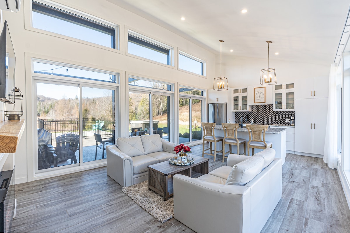 A modern open-concept living area features large oversized windows allowing natural light to fill the space. Comfortable white sofas are arranged around a wooden coffee table, with a dining area visible in the background that includes stylish bar stools and contemporary cabinetry.