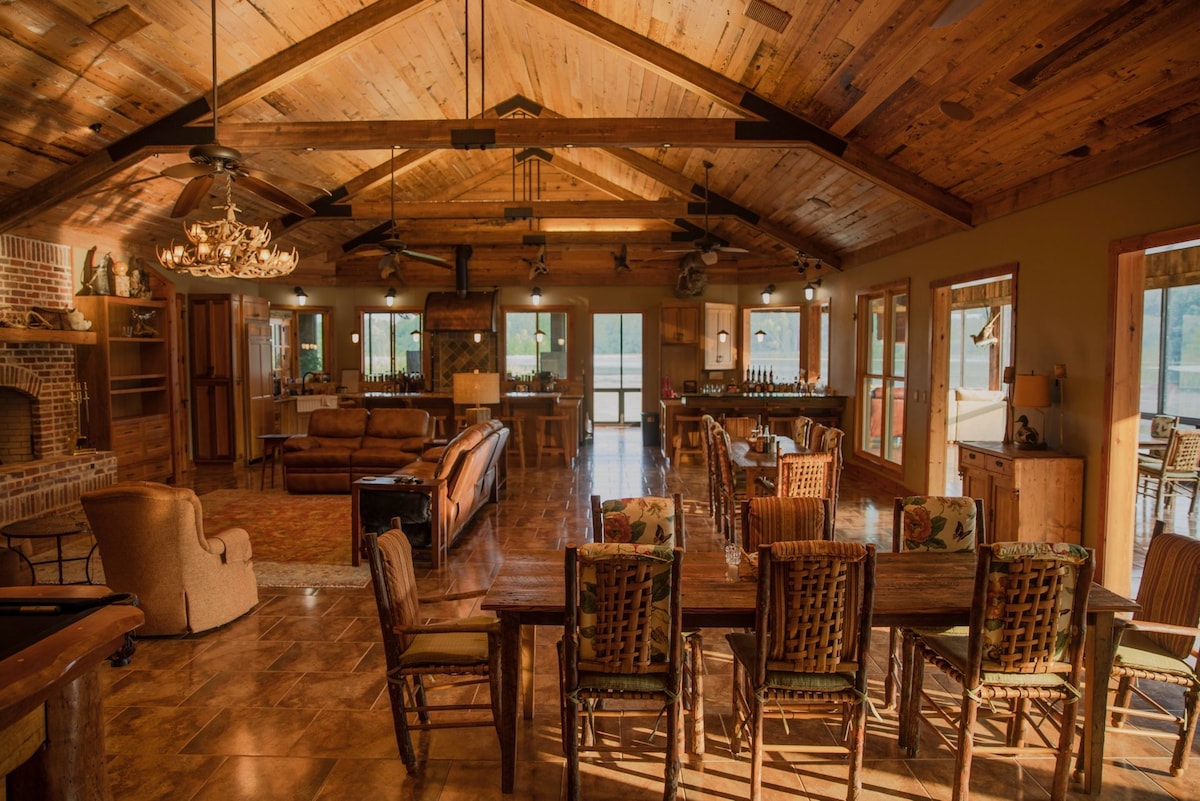 An expansive open-concept living and dining area features a high wooden ceiling with exposed beams. A large dining table surrounded by wicker chairs is positioned near multiple windows, allowing natural light to fill the space. Cozy seating arrangements include sofas and armchairs, enhancing the social atmosphere.