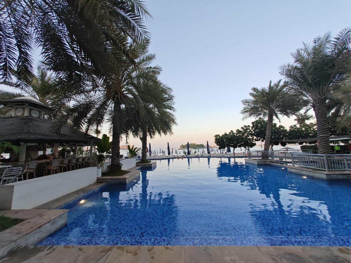 A tranquil outdoor pool is surrounded by palm trees, providing a relaxing atmosphere. Loungers line the water's edge, and a shaded cabana is visible on the left. In the background, the beach and ocean can be seen, completing the serene seaside setting.