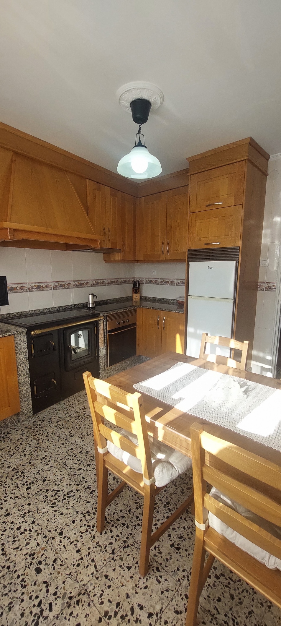 A functional kitchen is presented with wooden cabinets and a black countertop. Essential appliances, including a stove and refrigerator, are visible. A wooden dining table with four chairs occupies the center, illuminated by overhead lighting that enhances the light-colored walls.