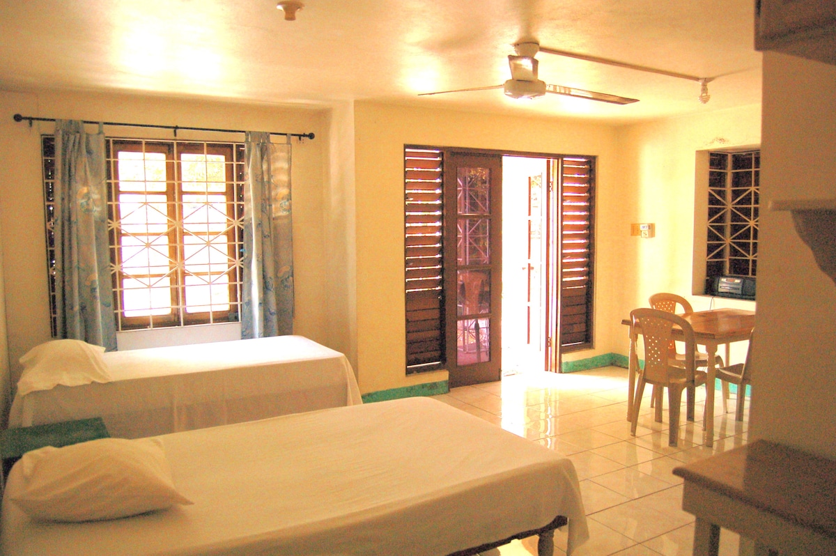 A spacious studio room is depicted with two single beds, light-coloured linens, and tiled flooring. Natural light enters through the vented wooden windows, accented by light curtains. A small dining table with plastic chairs is positioned near the entrance, creating a functional living space.