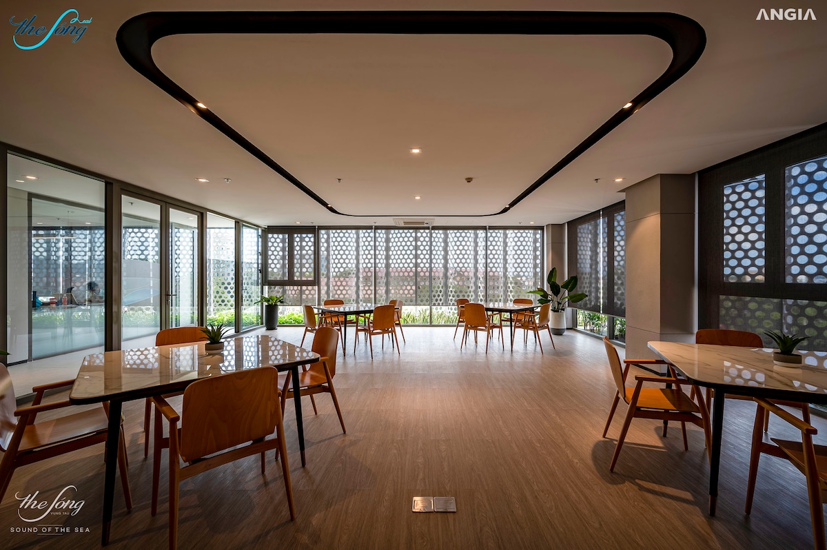 A modern dining area is presented with multiple wooden tables and chairs arranged neatly. Large windows allow natural light to illuminate the space, which is bordered by decorative screens and greenery visible outside, creating a welcoming atmosphere for communal meals.